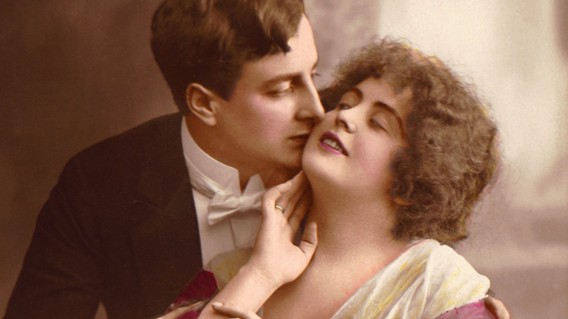 Hand tinted photograph shows a man in evening dress kisses a woman's cheek and embraces her, early 20th Century. (Photo by Stock Montage/Getty Images) - Credit: Getty Images