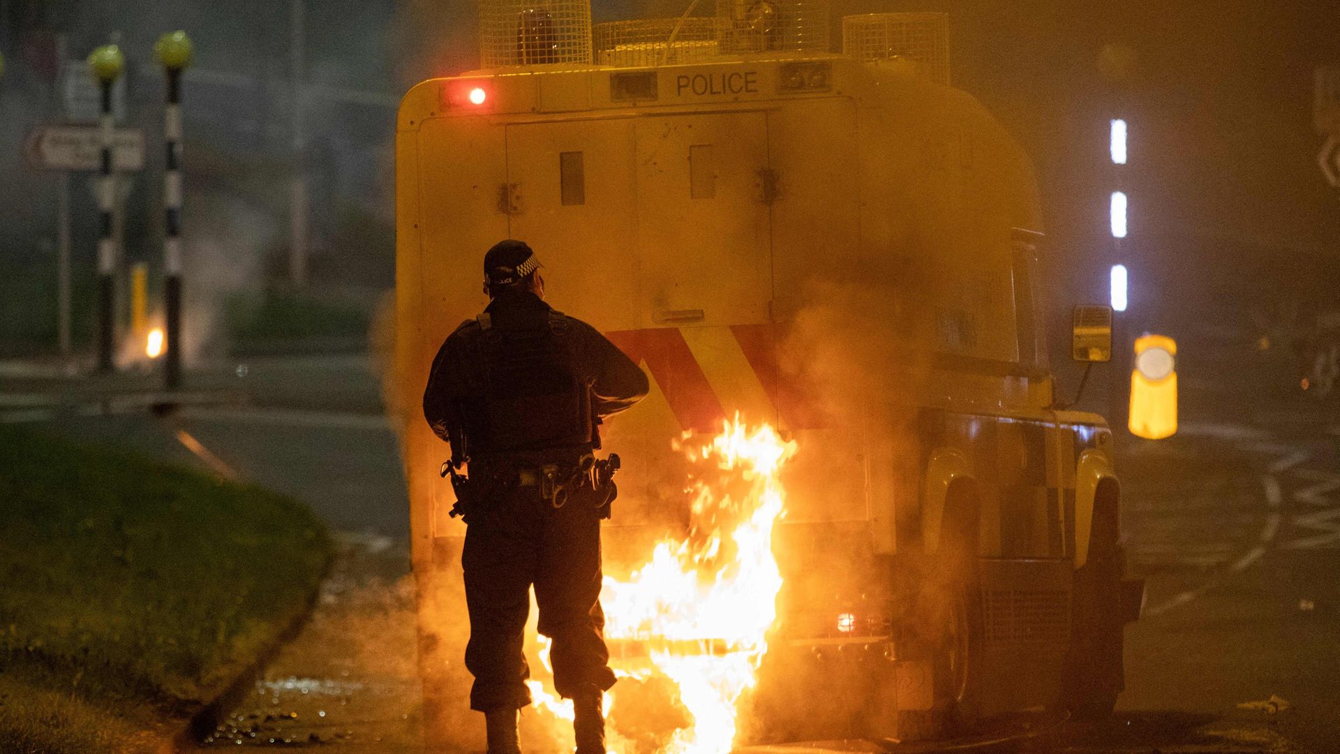 A police officer walks behind a police vehicle with flames leaping up the rear after violence broke out in Newtownabbey, north of Belfast, in Northern Ireland - Credit: Photo by PAUL FAITH/AFP via Getty Images