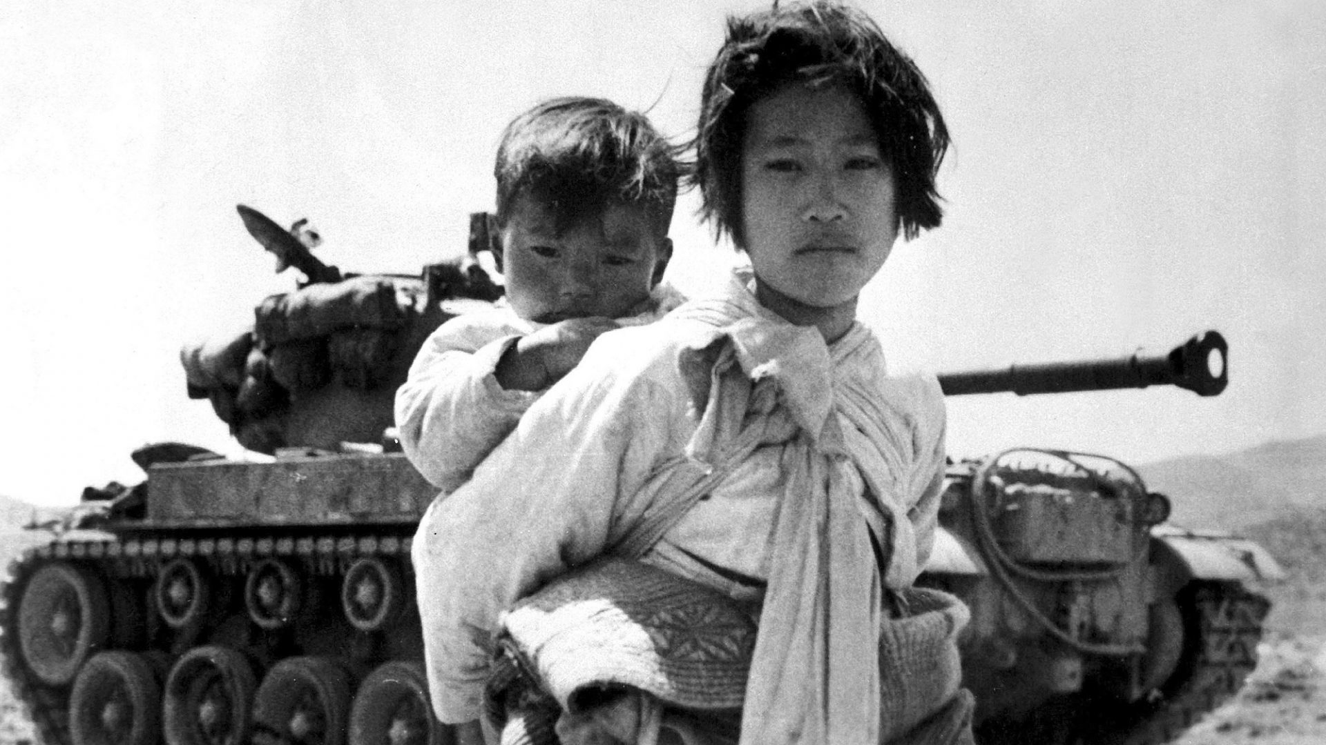 Carrying her baby brother on her back, a war-weary Korean girl walks by a stalled M-26 tank at Haengju, June 1951. Image: RV Spencer/Interim Archives/Getty