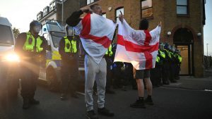 Protesters wrapped in the St George flag of England stand in front of a line of riot police as protesters march into the town centre of Epping. Photo: JUSTIN TALLIS/AFP via Getty Images