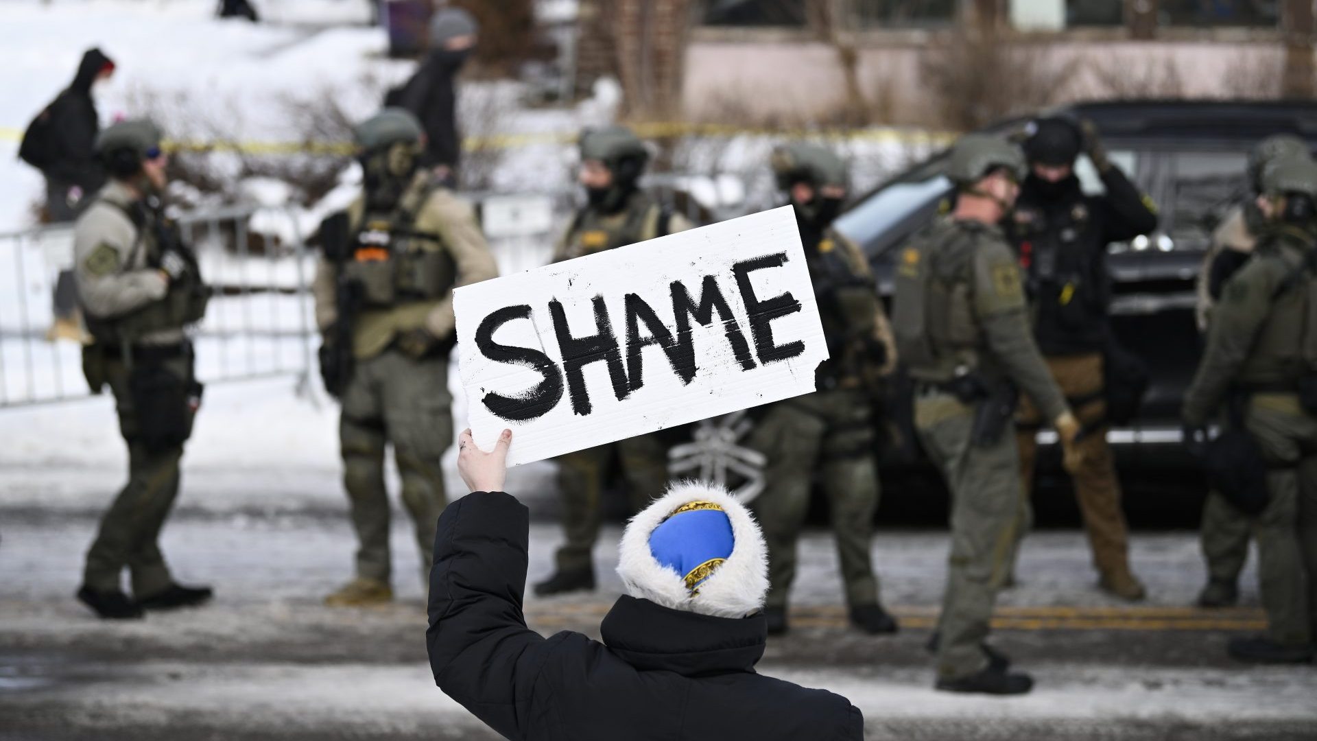 An onlooker holds a sign that reads "Shame" as members of law enforcement work the scene following a suspected shooting by an ICE agent during federal law enforcement operations.
