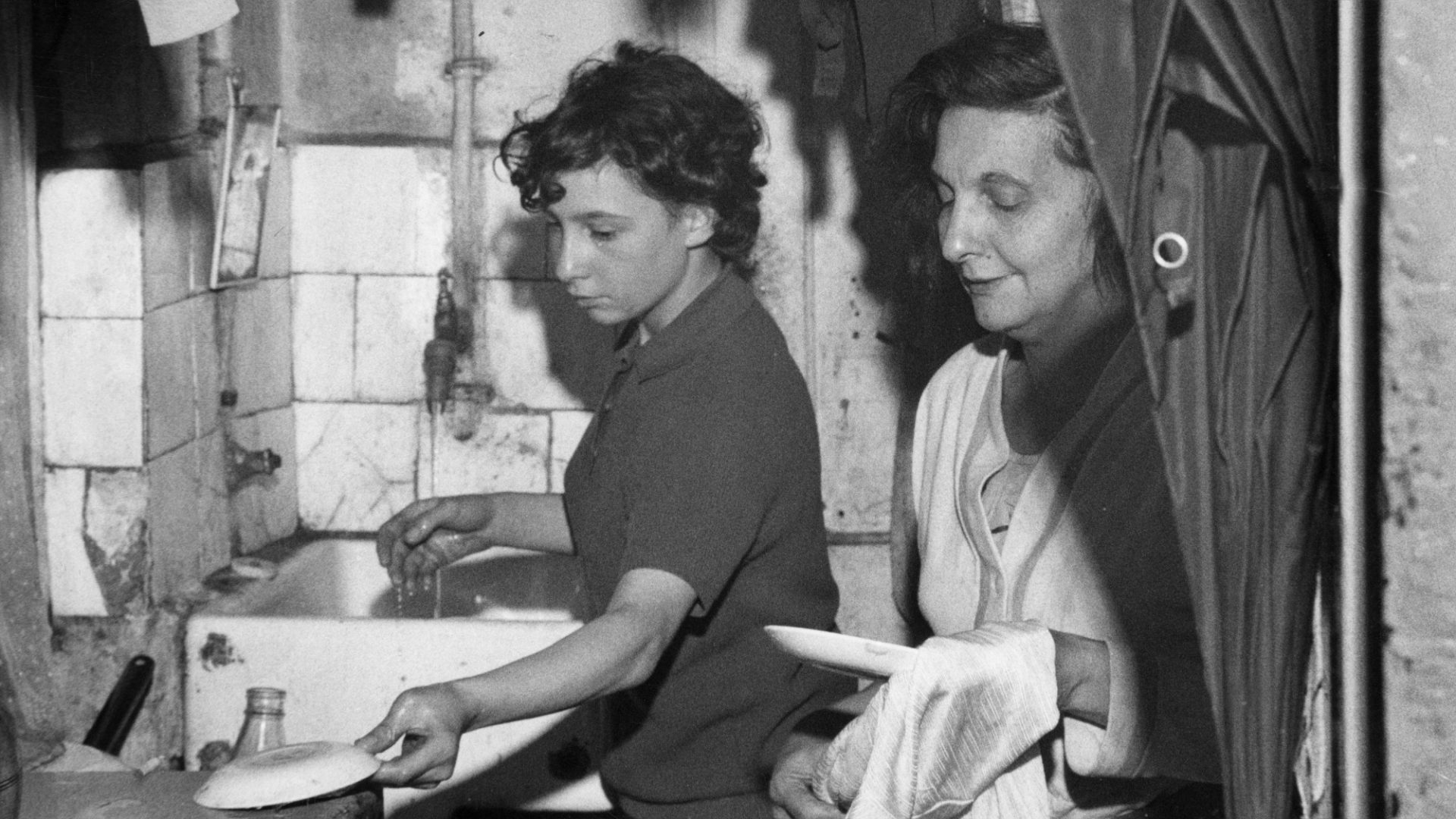 Women wash and dry plates in the cramped kitchen of a slum dwelling in London’s East End, 1965. Image: Jewish Chronicle/Heritage Images/Getty